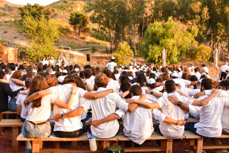 large group of teens in white sitting on benches and hanging on eachothers shoulders.