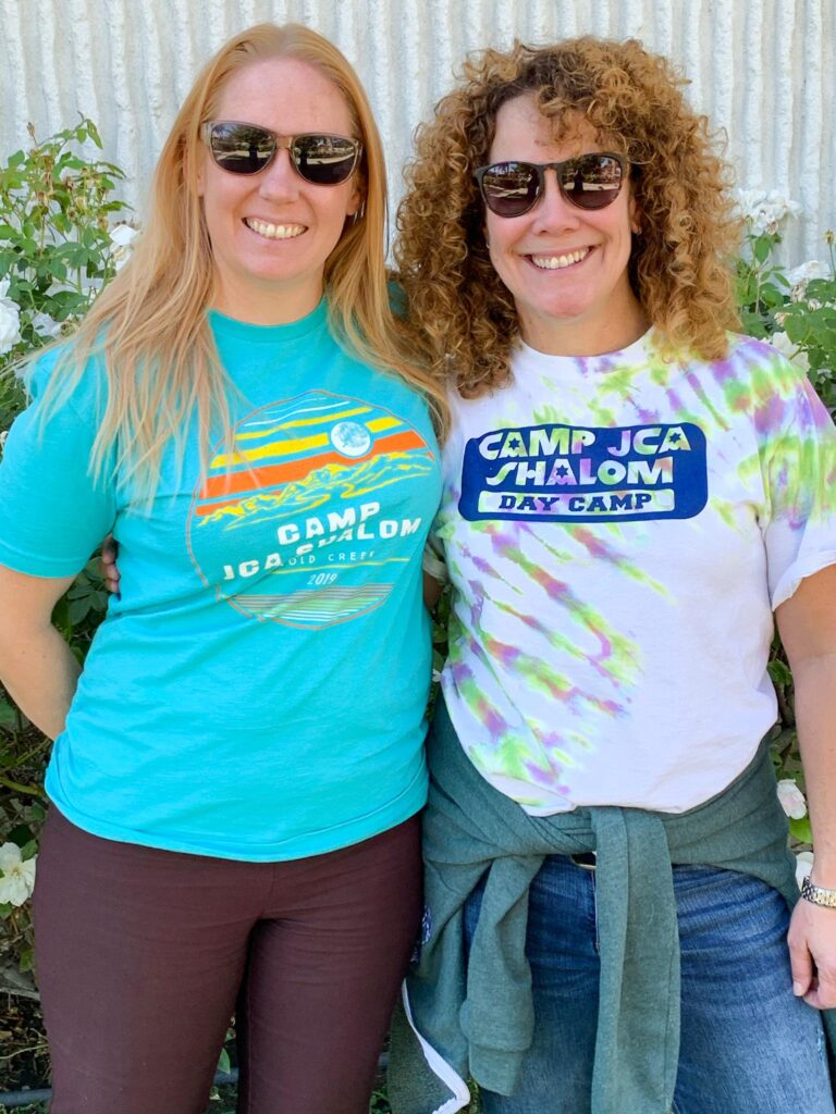 two women in camp jca shalom shirts smiling.