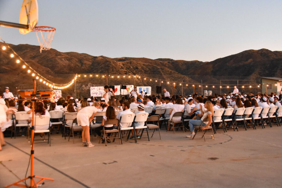 large group of teens sitting in white folding chairs outside on a basketball court.